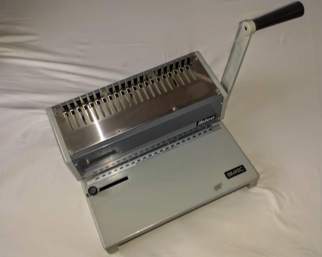 Desktop comb binding machine with a punch lever and an open comb spine, loaded with letter-size paper on an office desk