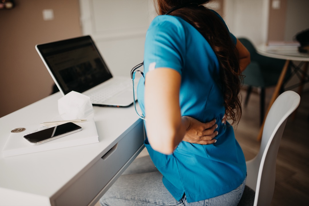 Office worker pressing a hand to the lower back while seated at a desk workstation, the posture problem a lumbar support pillow is bought to fix