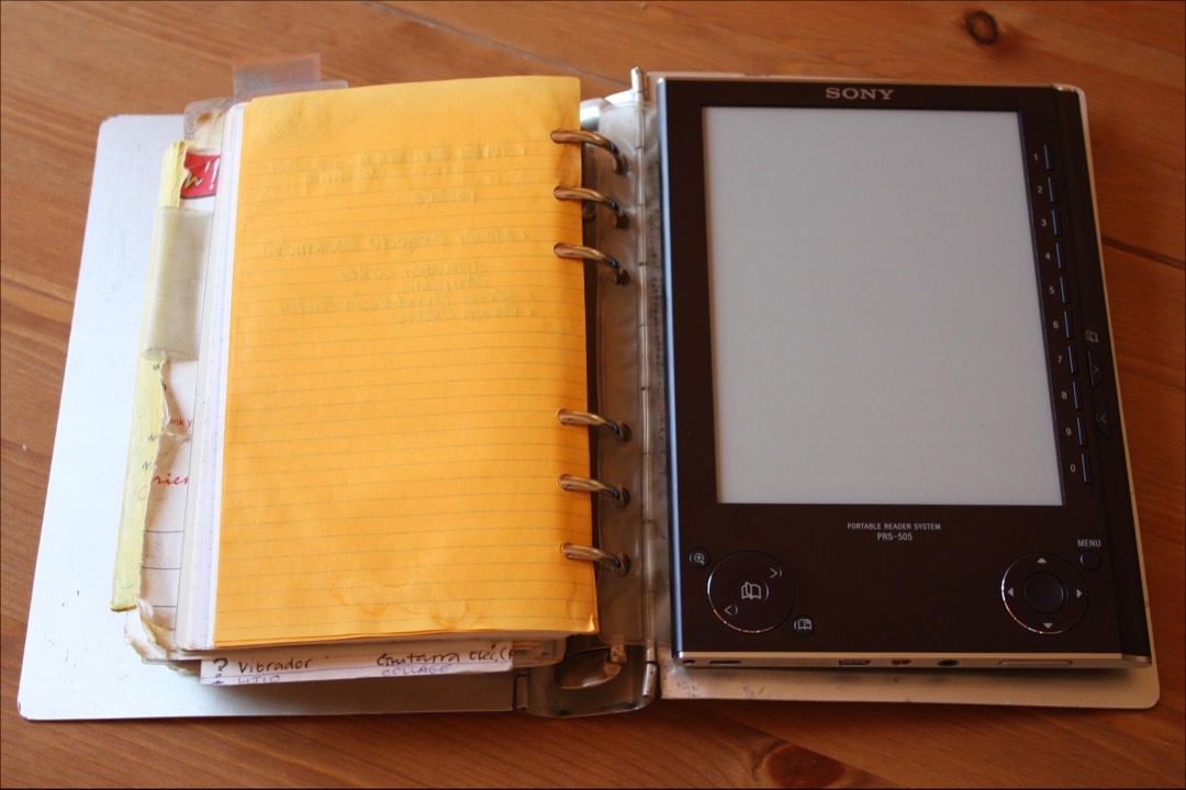 An open paper planner on a desk beside a digital e-reader device