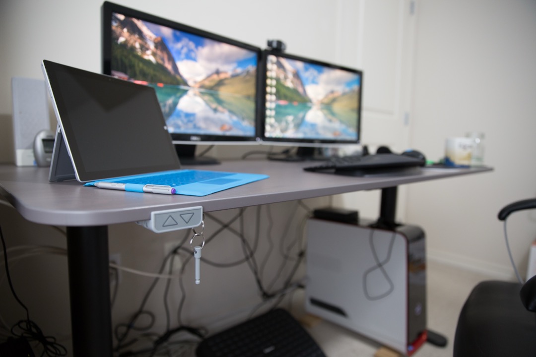 A height-adjustable sit-stand desk with dual monitors, a laptop, and a visible up-down height control paddle mounted under the desktop
