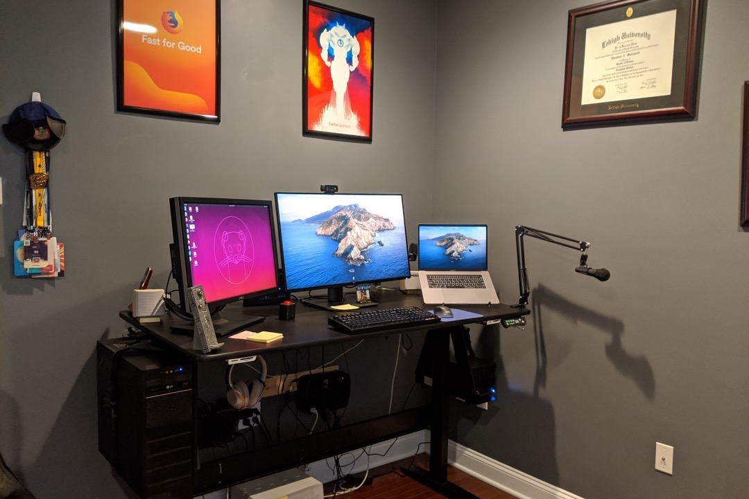 A height-adjustable desk with dual monitors and a docked laptop serving as a third screen in a home office