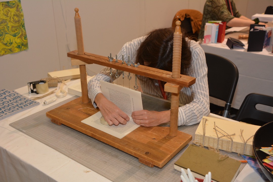A person sewing a book's signatures on a wooden bookbinding sewing frame, with a finished sewn text block showing exposed cords resting beside it