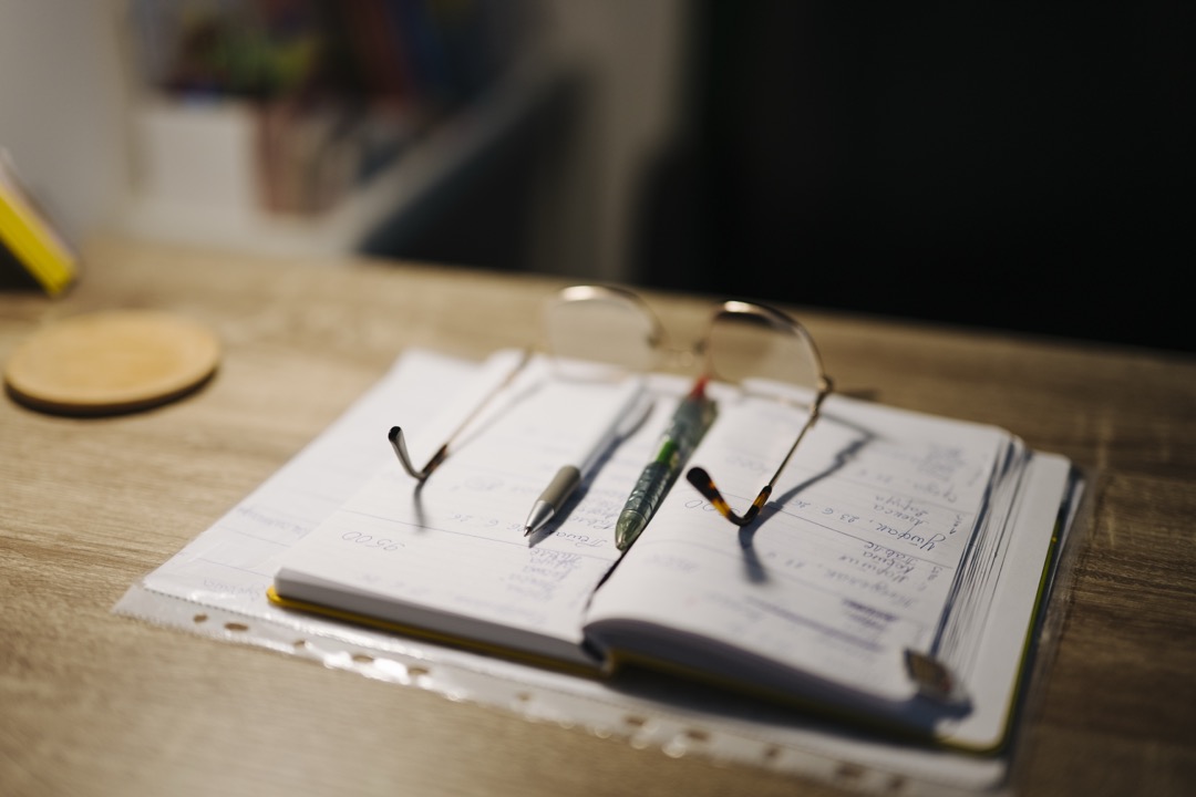 An open handwritten notebook with two pens and a pair of reading glasses on a wooden desk