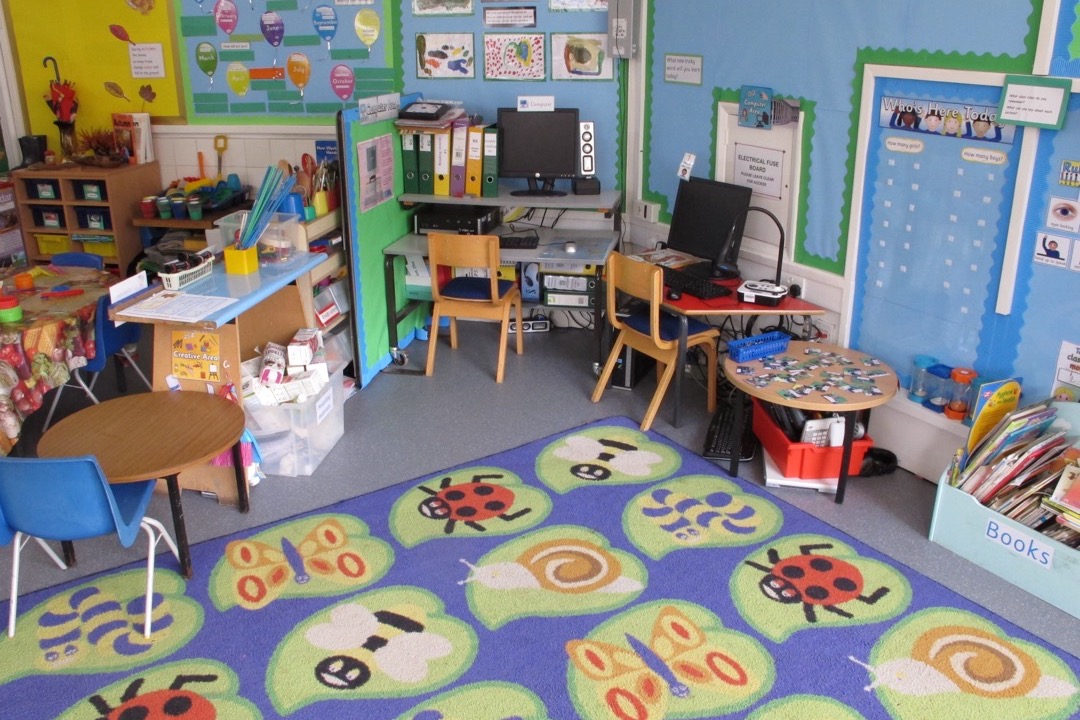 A brightly lit elementary classroom with student desks in clusters, labeled supply bins, wall displays, and a whiteboard at the front