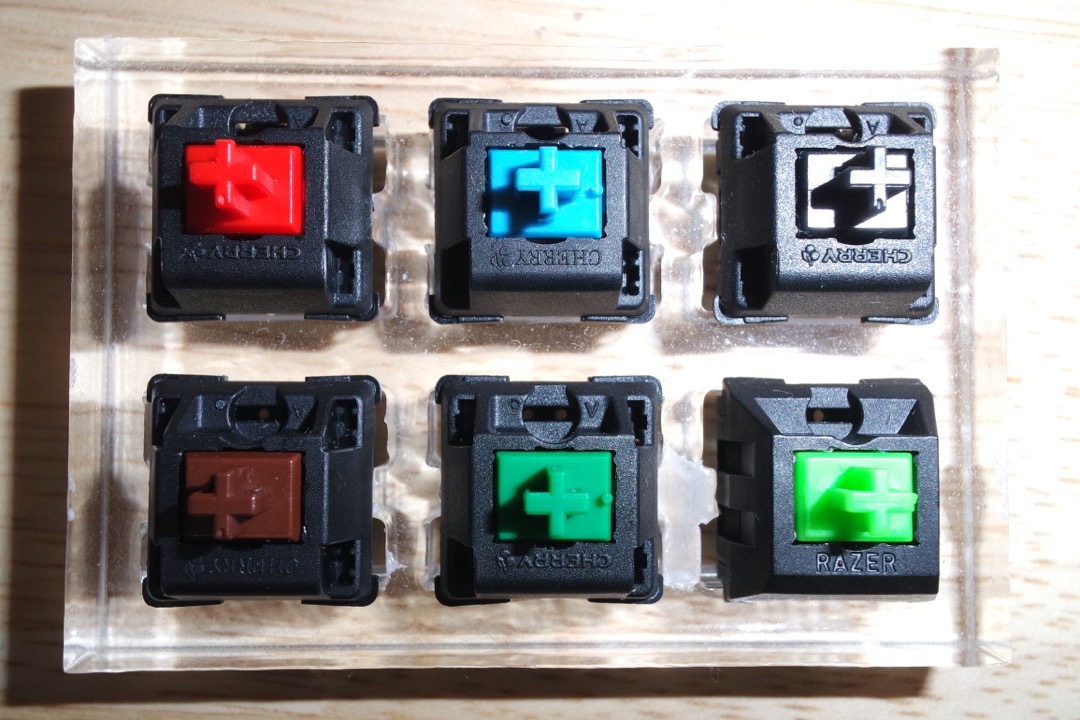 A clear switch tester holding six mechanical keyboard switches — Cherry MX red, blue, clear, brown, green, and a Razer green