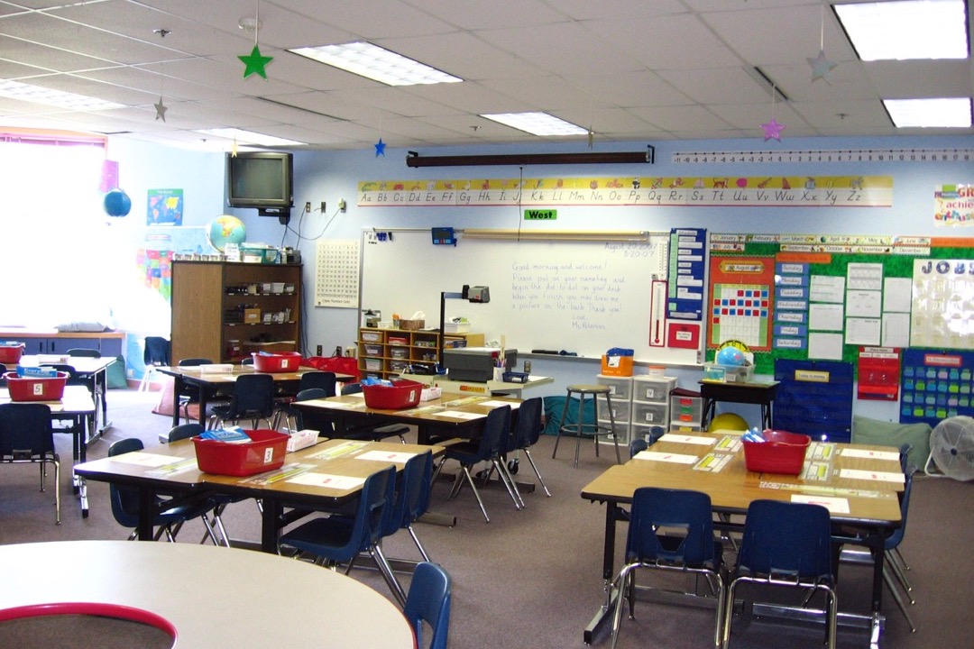 A set-up elementary classroom with student desks arranged in groups, a whiteboard, anchor charts on the walls, labeled supply bins, and a reading corner